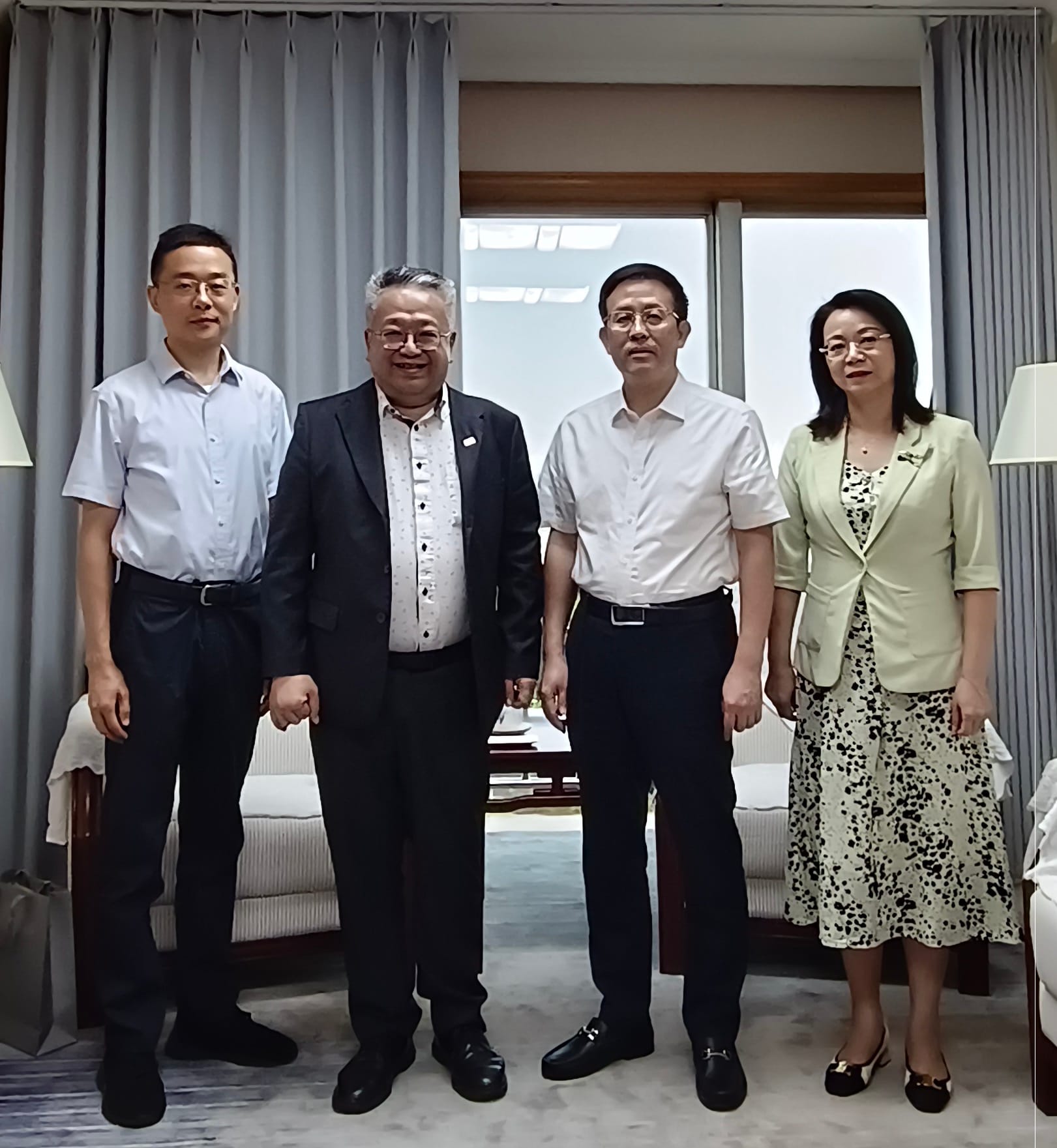 Four people in business attire pose indoors near a window with curtains.